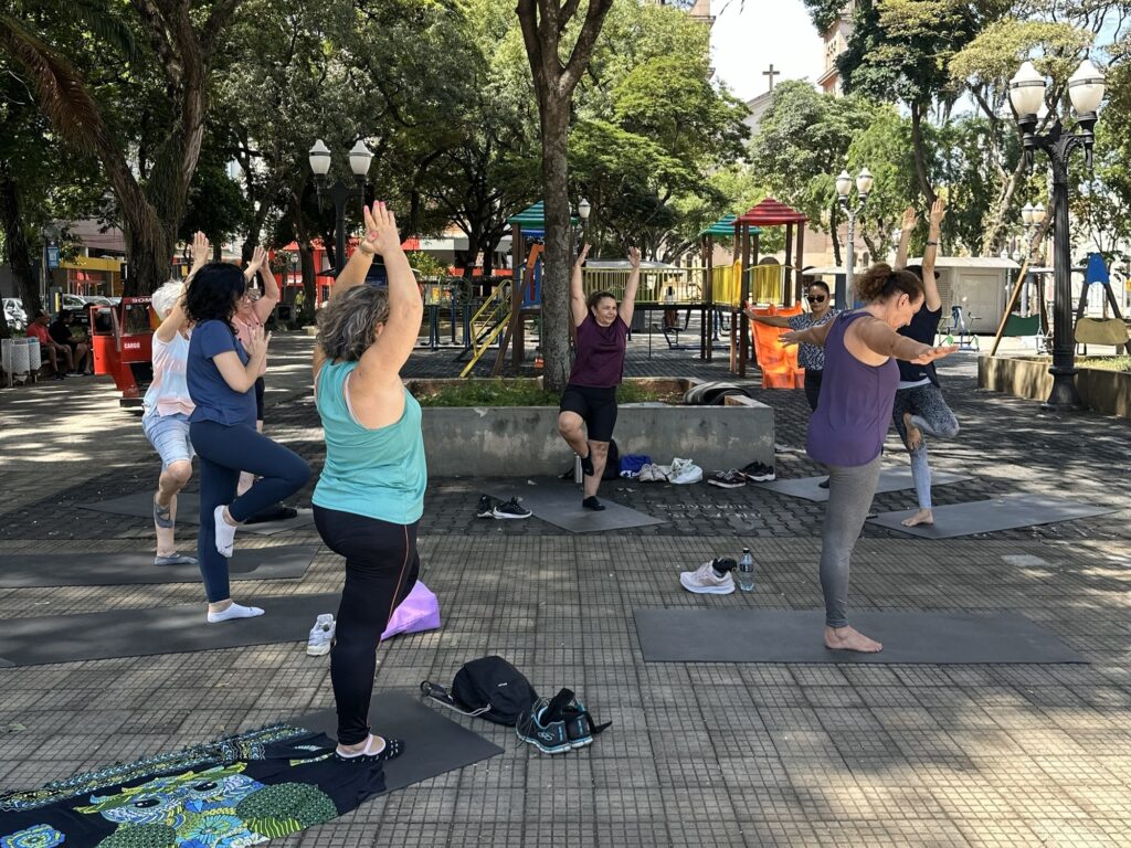 A imagem mostra um grupo de mulheres praticando yoga ao ar livre em um parque. Elas estão em diferentes posições de yoga, com os braços levantados e um pé apoiado no chão. O parque tem árvores, bancos e um parquinho infantil ao fundo. O tempo está ensolarado e as pessoas estão vestindo roupas confortáveis para a prática de exercícios.