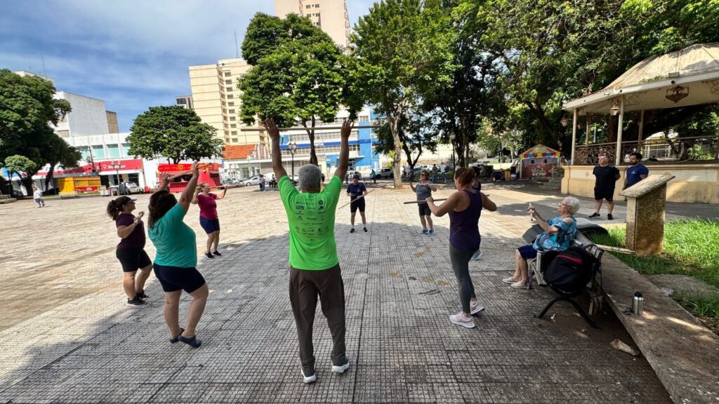A imagem mostra um grupo de pessoas praticando exercícios ao ar livre em uma praça. No centro da imagem, um homem mais velho, vestindo uma camiseta verde e calças marrons, está de costas para a câmera, com os braços erguidos segurando um bastão. Ao redor dele, outras pessoas, a maioria mulheres, também estão com bastões, realizando movimentos de alongamento ou exercício. Mais ao fundo, em uma área com árvores e um coreto, observa-se um grupo de pessoas conversando, e uma senhora idosa sentada em uma cadeira de rodas, parecendo participar ou observar a atividade. Há edifícios ao fundo, indicando que a praça está localizada em uma área urbana. O dia parece ensolarado, com céu azul e nuvens.