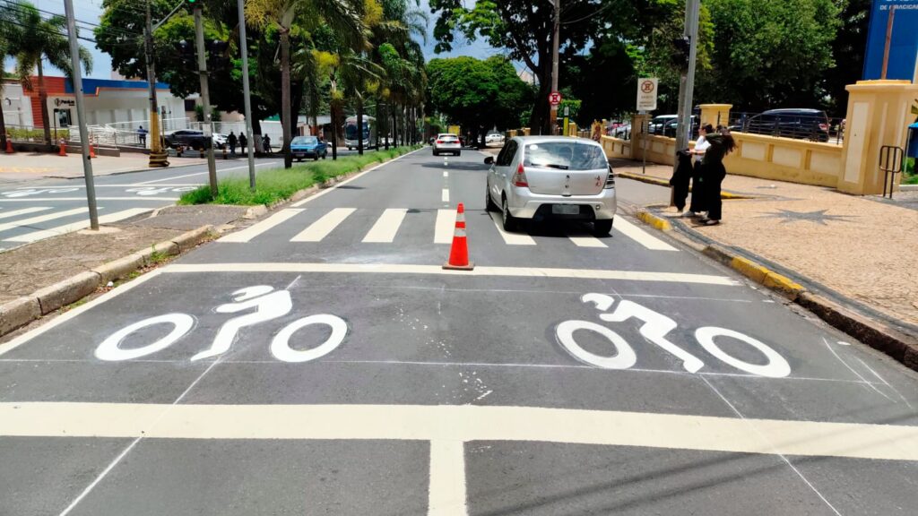 A imagem mostra uma rua com uma ciclovia recém-pintada. A ciclovia é marcada com símbolos de bicicleta em branco sobre o asfalto escuro. Há também faixas de pedestres brancas atravessando a rua. Um cone de trânsito laranja está posicionado em uma das faixas de pedestres. Um carro prateado está parado em uma faixa de pedestres, e outros carros podem ser vistos ao fundo, juntamente com árvores e edifícios. Pessoas estão na calçada, e a ciclovia parece estar em uma área urbana.