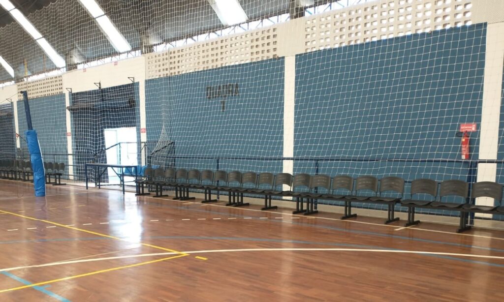 A imagem mostra o interior de um ginásio poliesportivo. O chão é de madeira, com marcações de quadra em diferentes cores (amarelo, azul e branco). Ao fundo, há uma parede azul com uma rede de proteção pendurada, dividindo o espaço. Há uma fileira de assentos pretos para espectadores ao longo da parede. À esquerda, um poste azul acolchoado indica que pode ser uma estrutura para rede (como de vôlei ou tênis). Há também uma porta aberta ao fundo, por onde se pode ver o exterior. O teto é alto, com iluminação artificial embutida e uma estrutura de treliça metálica. Uma placa vermelha com a palavra "EXTINTOR" está visível na parede, indicando a presença de um extintor de incêndio.