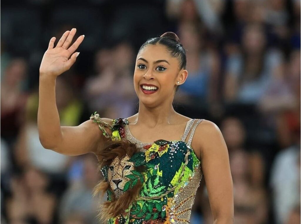A imagem mostra uma ginasta rítmica sorrindo e acenando para a câmera. Ela está usando um maiô vibrante e detalhado, adornado com um leão estilizado no peito, folhas verdes e pedrarias brilhantes. Seu cabelo está preso em um coque elegante, e ela tem uma maquiagem marcante com delineador nos olhos. O fundo está desfocado, sugerindo que ela está em uma arena ou ginásio com público.
