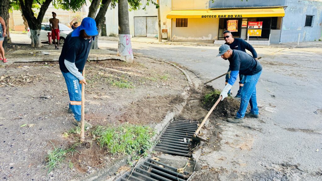 A imagem mostra três trabalhadores realizando a limpeza e manutenção de um bueiro em uma rua urbana, utilizando ferramentas manuais para remover terra e vegetação acumulada ao redor da grade de drenagem. Eles estão uniformizados e equipados com luvas e chapéus de proteção, enquanto ao fundo aparecem algumas pessoas próximas a um carro estacionado e um bar com toldo amarelo.
