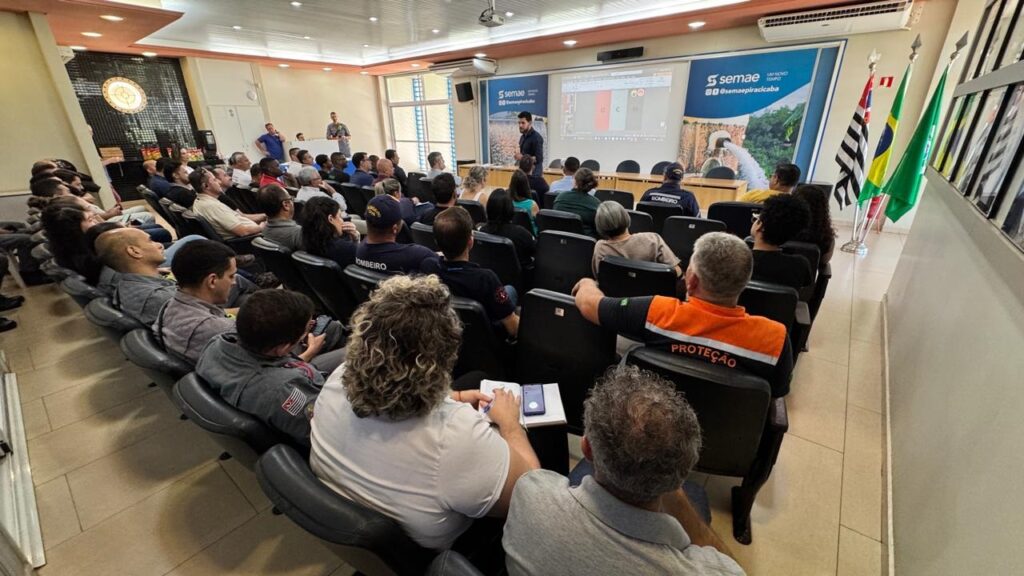 A imagem mostra uma sala de treinamento ou conferência com uma plateia sentada em cadeiras, voltada para uma tela de projeção. Há duas pessoas em pé na frente da sala, uma delas apresentando algo na tela. A plateia parece ser composta principalmente por homens, muitos dos quais vestem uniformes, possivelmente de bombeiros ou de alguma organização de segurança, indicados pelas palavras "BOMBEIRO" e "PROTEÇÃO" visíveis em algumas vestimentas. O ambiente é iluminado por luzes no teto e há bandeiras visíveis no lado direito da sala, sugerindo um evento oficial ou institucional. A tela de projeção exibe um slide com gráficos e texto, e ao lado dela, há um logotipo com a palavra "semae". A atmosfera geral sugere uma apresentação informativa ou treinamento.