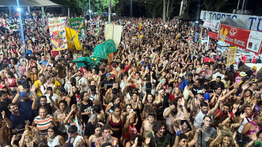 A imagem retrata uma multidão vibrante e animada durante um evento noturno, possivelmente um carnaval ou festival. A maioria das pessoas está em pé, celebrando e interagindo, com muitas mãos erguidas em sinal de alegria e participação. A iluminação artificial, proveniente de postes e talvez de barracas, cria uma atmosfera festiva, destacando os rostos e as fantasias coloridas. Ao fundo, observam-se tendas e estruturas temporárias, indicando a presença de vendedores de comida e bebida, como sugerem as placas com os dizeres "BARRACA DO PASTEL" e "LANCHE PERALDVIND". Há também faixas e banners coloridos, alguns com mensagens em português como "JUNTES, EM MISTURAES PRESERVANDO NOSSO". A presença de ornamentos e adereços carnavalescos, como confetes e máscaras, é notável em alguns indivíduos, reforçando a ideia de celebração. A atmosfera geral é de euforia e união, com as pessoas imersas na energia do evento.