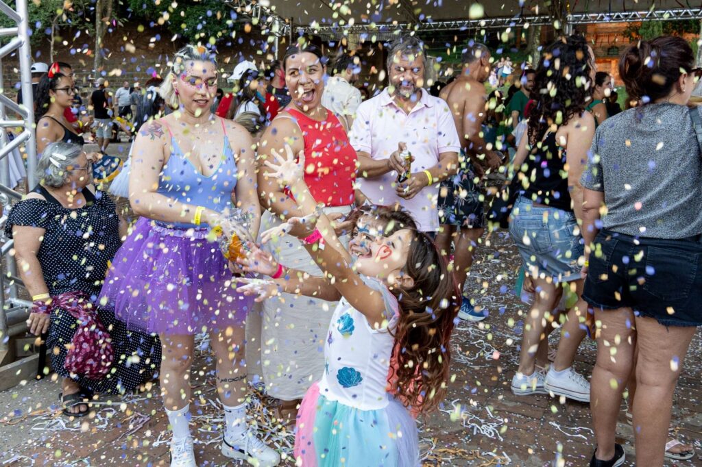 A imagem mostra um grupo de pessoas em uma celebração ao ar livre, possivelmente um carnaval ou festival, com confetes coloridos caindo sobre elas. No centro, uma menina com um vestido colorido e maquiagem no rosto, com os braços levantados, parece estar se divertindo com a chuva de confetes. Ao redor dela, outras pessoas, incluindo mulheres e homens, também estão participando da festa, com expressões de alegria e surpresa. Uma mulher à esquerda usa um vestido azul claro e uma saia roxa de tule, enquanto outra, à sua direita, veste uma blusa vermelha. Um homem ao fundo usa uma camisa rosa. A atmosfera geral é de celebração e alegria.