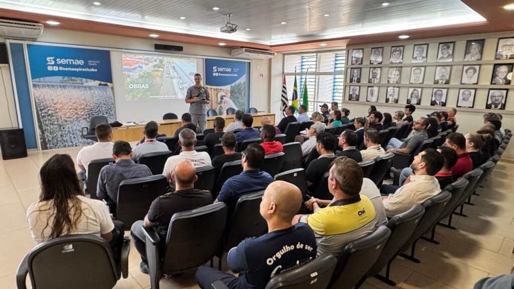 A imagem mostra uma sala de reuniões onde um homem, vestindo um uniforme cinza, está em pé em frente a uma tela de projeção e fala para uma audiência sentada em cadeiras. A tela exibe o logotipo da "Semae" e uma imagem de obras. As paredes da sala são adornadas com uma série de retratos emoldurados, possivelmente de personalidades importantes ou ex-diretores da organização. A audiência é composta por homens e algumas mulheres, a maioria vestindo roupas casuais. A atmosfera parece ser de uma apresentação ou reunião informativa.