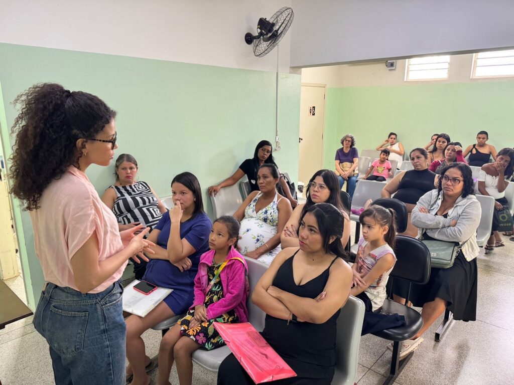 A imagem mostra um grupo de mulheres sentadas em cadeiras em uma sala com paredes verdes claras. Uma mulher com cabelo cacheado escuro e óculos está de pé, de frente para o grupo, gesticulando com as mãos enquanto fala. Ela usa uma blusa rosa claro e jeans. As mulheres sentadas parecem estar prestando atenção à palestrante. Há uma mulher grávida sentada no meio da sala. Algumas das mulheres estão com os braços cruzados, e outras parecem pensativas. Há também algumas crianças pequenas sentadas entre as mulheres. Ao fundo, há um ventilador de teto preto preso à parede e algumas janelas com venezianas. Há também portas e um quadro de avisos com símbolos. A iluminação na sala parece ser artificial, vinda de luminárias no teto. A atmosfera geral sugere uma reunião informativa ou uma aula, possivelmente relacionada à saúde materna ou familiar.