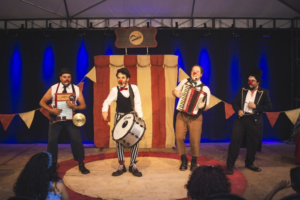 A imagem mostra quatro palhaços no palco, apresentando-se para uma audiência. Cada palhaço tem uma maquiagem distinta, com narizes vermelhos e expressões faciais exageradas. Eles usam figurinos coloridos e variados, com chapéus, coletes e calças listradas. Os palhaços estão tocando instrumentos musicais: um toca um gaita de foles, outro um tambor, um terceiro um acordeão e o quarto um triângulo. O palco está decorado com uma cortina vermelha e branca no fundo, bandeiras coloridas e iluminação azul que cria um ambiente festivo. Há também uma plateia visível na parte inferior da imagem, sugerindo que a performance está em andamento. A atmosfera geral é de alegria e entretenimento.