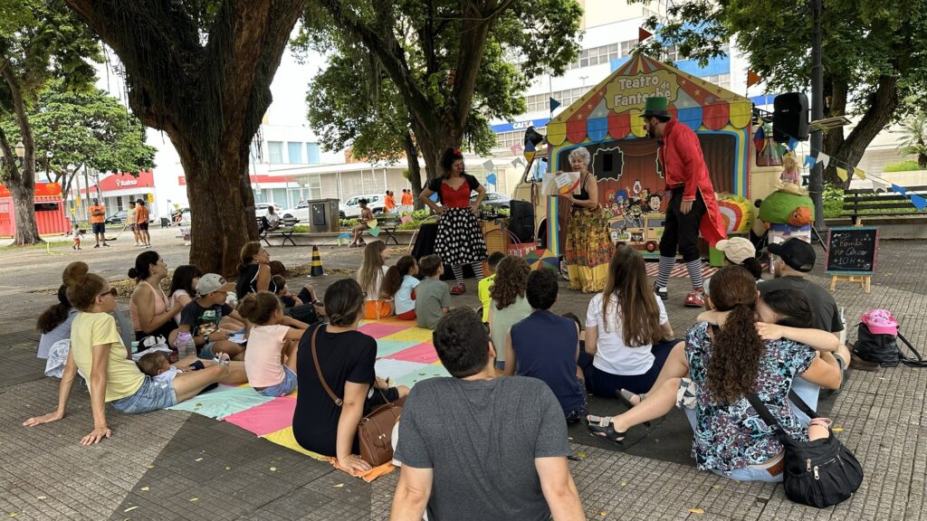 A imagem mostra uma cena ao ar livre, possivelmente um parque ou praça, onde um grupo de pessoas, incluindo adultos e crianças, está reunido para assistir a uma apresentação de teatro. Ao fundo, há um pequeno palco colorido com a inscrição "Teatro de Fantoche", decorado com bandeirinhas. No palco, três artistas estão em destaque. Uma mulher com um vestido estampado e um xale colorido segura um livro, parecendo narrar uma história. Ao seu lado, um homem vestindo um casaco vermelho vibrante, calças pretas e meias listradas com um chapéu verde, está em uma pose que sugere atuação. Outra mulher, com um vestido vermelho e uma saia de bolinhas pretas, está de pé com as mãos na cintura, possivelmente como uma personagem ou apresentadora. A audiência está sentada em um tapete colorido no chão, com as costas voltadas para a câmera em sua maioria. As crianças estão sentadas na frente, atentamente observando a performance, enquanto os adultos estão atrás delas. Há árvores frondosas ao redor, proporcionando sombra, e edifícios ao fundo indicam que a cena se passa em uma área urbana. A atmosfera geral é de diversão e envolvimento familiar.