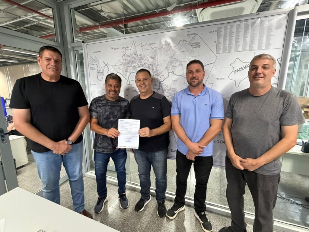 A imagem mostra cinco homens em pé em um escritório, com um grande mapa ao fundo. Os homens estão vestidos casualmente, a maioria com camisetas e jeans. O homem mais à esquerda usa uma camiseta preta e jeans claros, com um relógio no pulso esquerdo. O segundo homem da esquerda para a direita veste uma camiseta com estampa tie-dye e jeans escuros. Ele segura um documento com as duas mãos. O homem do meio usa uma camiseta preta e jeans escuros. O quarto homem da esquerda para a direita veste uma polo azul clara e calças pretas. O homem mais à direita veste uma camiseta cinza e calças cinzas escuras. Todos os homens sorriem para a câmera. O mapa atrás deles parece ser um mapa urbano ou regional, com nomes de bairros ou áreas, como "FAZ CACHOEIRA", "SANTA LIDIA", "DISTRITO INDUSTRIAL UNINORTE" e "IRACEMÁ". Há também rios ou corpos d'água representados no mapa. O escritório parece moderno, com teto aparente com tubulações e luminárias, e grandes janelas. A iluminação é clara, sugerindo um ambiente de trabalho.