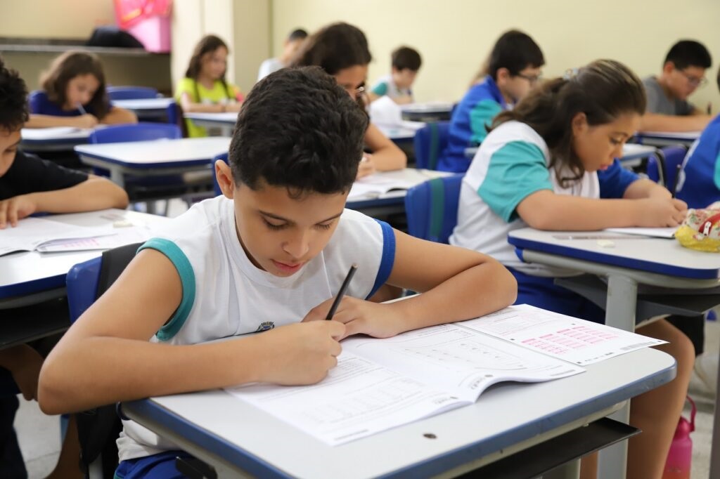 A imagem mostra uma sala de aula com vários alunos sentados em carteiras, concentrados em suas tarefas. No primeiro plano, um menino com uma camiseta branca e detalhes azuis está escrevendo em um caderno com um lápis. Ele parece estar fazendo uma prova ou um exercício. Ao fundo, outros alunos também estão escrevendo ou lendo. As carteiras são organizadas em fileiras, e a iluminação da sala sugere que seja durante o dia. A atmosfera geral é de concentração e estudo.