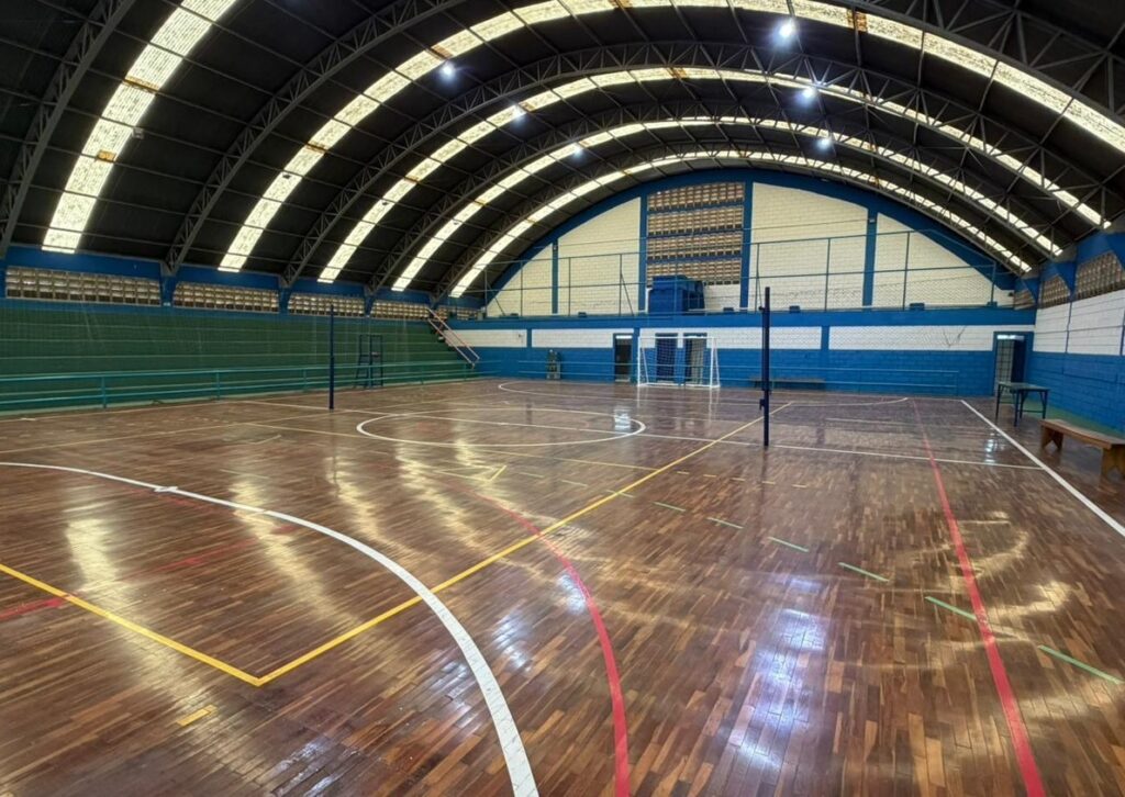 A imagem mostra o interior de uma quadra poliesportiva coberta. O piso é de madeira com marcações coloridas para diferentes esportes, como basquete e vôlei. O teto é arqueado e sustentado por uma estrutura metálica escura, com painéis translúcidos que permitem a entrada de luz natural, complementada por iluminação artificial. As paredes são pintadas em tons de azul e branco, com áreas em verde que parecem ser arquibancadas ou barreiras. Na quadra, há equipamentos esportivos, como postes de rede de vôlei e uma pequena baliza de futebol. A iluminação do piso de madeira reflete a luz, dando um aspecto brilhante e bem conservado ao local. Ao fundo, observa-se uma porta e uma janela com grades.