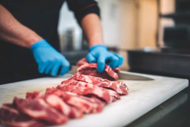 A imagem mostra em close-up as mãos de uma pessoa usando luvas azuis, cortando pedaços de carne crua em uma tábua de corte branca. A carne está disposta em uma fila na tábua, com os cortes frescos e vermelhos. O fundo está desfocado, mas sugere um ambiente de cozinha profissional. A iluminação destaca a textura da carne e o brilho das luvas.