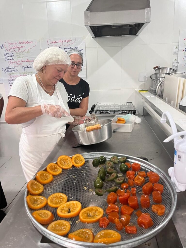 A imagem mostra uma cozinha comercial onde duas mulheres estão preparando frutas cristalizadas ou desidratadas. Em primeiro plano, há uma bandeja grande e circular com uma tela metálica, contendo fatias de laranja dispostas em um dos lados, figos verdes inteiros e cortados ao meio no centro, e cubos de abóbora ou mamão em um dos lados. Uma das mulheres, vestindo touca, avental branco e luvas, está usando uma escumadeira para retirar pedaços de uma panela grande. Ao fundo, outra mulher com touca e óculos observa o processo. A cozinha tem paredes azulejadas brancas, bancada de aço inox, fogão a gás e coifa. Na parede, há cartazes escritos à mão em português que explicam processos de desidratação e cristalização de frutas, listando ingredientes como laranja, abacaxi e abóbora. A iluminação é clara, típica de ambiente de trabalho. A imagem transmite um ambiente de produção artesanal ou semi-industrial de frutas cristalizadas.