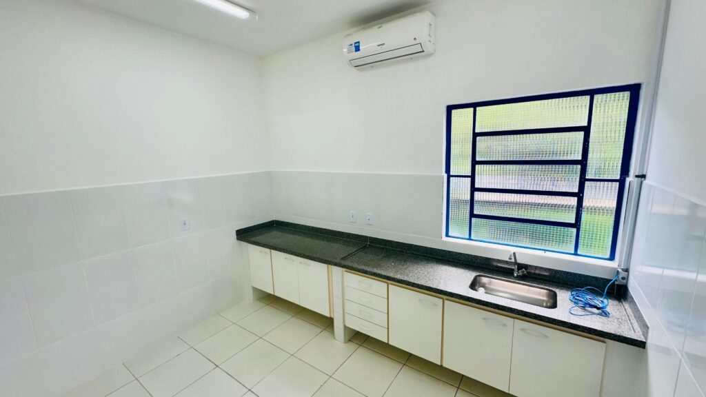 A imagem mostra uma cozinha pequena e funcional. * As paredes são revestidas com azulejos brancos até a metade da altura, e a parte superior é pintada de branco. * Há um balcão de granito escuro que se estende ao longo de uma das paredes, com uma pia de inox instalada. * Abaixo do balcão, há armários brancos com puxadores discretos e gavetas com frentes em tom amadeirado claro. * Uma janela com grades azuis e vidro texturizado permite a entrada de luz natural. * Um aparelho de ar condicionado está instalado na parede, acima do balcão. * O piso é revestido com azulejos quadrados de cor clara. * Há tomadas elétricas visíveis na parede acima do balcão. * Um rolo de fio azul está sobre o balcão, perto da pia.