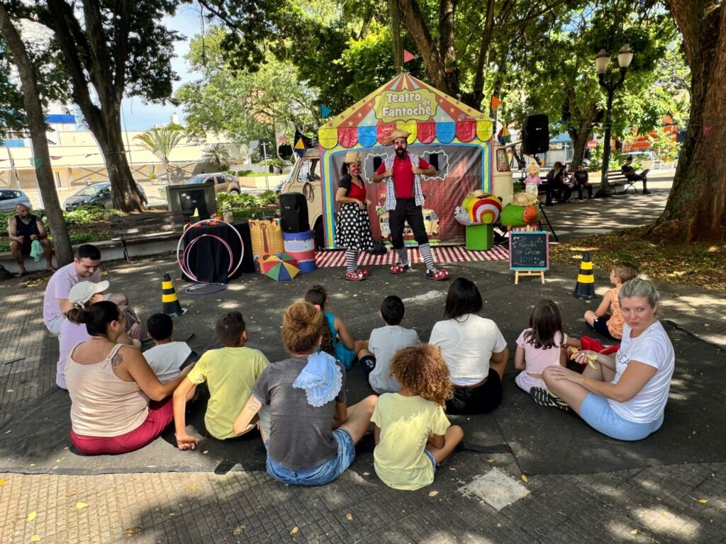 A imagem retrata uma cena vibrante de um show de fantoches ao ar livre, possivelmente em um parque ou praça, com árvores frondosas ao fundo. No centro, um palco colorido com a inscrição "Teatro de Fantoche" serve de cenário para dois artistas caracterizados como palhaços, com maquiagem exagerada, chapéus e roupas chamativas. Um deles, um homem com um colete xadrez aberto sobre uma camisa vermelha, faz gestos expressivos, enquanto uma mulher ao seu lado, vestida com um traje de bolinhas pretas e brancas, observa. A plateia, composta majoritariamente por crianças e alguns adultos, senta-se em um tapete preto no chão, voltada para o palco. As crianças, com idades variadas, demonstram atenção e curiosidade, algumas sentadas em grupo, outras mais próximas do palco. Ao redor do palco, há adereços como bambolês, tambores decorados, um guarda-chuva colorido e um personagem de lesma com um globo ocular grande, que adicionam um toque lúdico à apresentação. Uma pequena lousa com a inscrição "Kombi no Circo 10h" sugere o tema do espetáculo ou o horário de início. A atmosfera geral é de alegria e entretenimento familiar, com a luz do sol filtrando pelas árvores, criando um ambiente acolhedor.