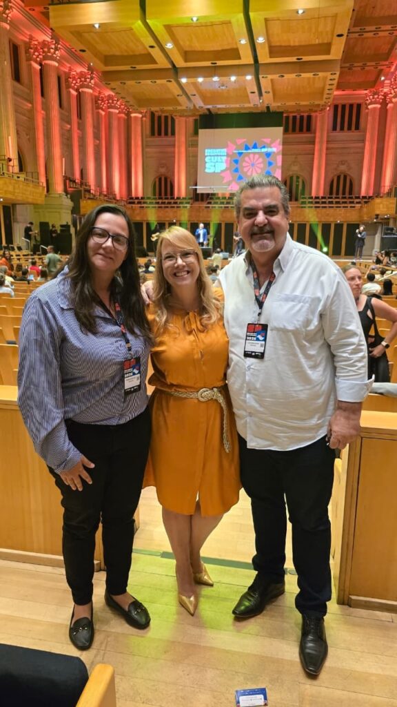 A imagem mostra três pessoas em pé, sorrindo e posando para uma foto em um evento. A mulher à esquerda usa um vestido listrado azul e branco, calças pretas e mocassins pretos. Ela usa óculos e tem cabelos escuros. A mulher do meio usa um vestido laranja vibrante com um cinto dourado e saltos dourados. Ela também usa óculos e tem cabelos loiros. O homem à direita usa uma camisa branca de botões, calças pretas e sapatos pretos. Ele tem cabelos escuros e grisalhos e barba. Todos os três estão usando crachás. O fundo da imagem sugere que eles estão dentro de um grande salão, possivelmente uma sala de concertos ou um centro de convenções. Há colunas altas e ornamentadas iluminadas com luzes laranjas quentes à esquerda e ao centro. Acima, o teto é de madeira com detalhes embutidos. Um grande banner com um design geométrico colorido e texto em branco e azul está visível no palco ao fundo. Há assentos em fileiras ao fundo e algumas pessoas estão sentadas neles. A iluminação geral é quente e convidativa.