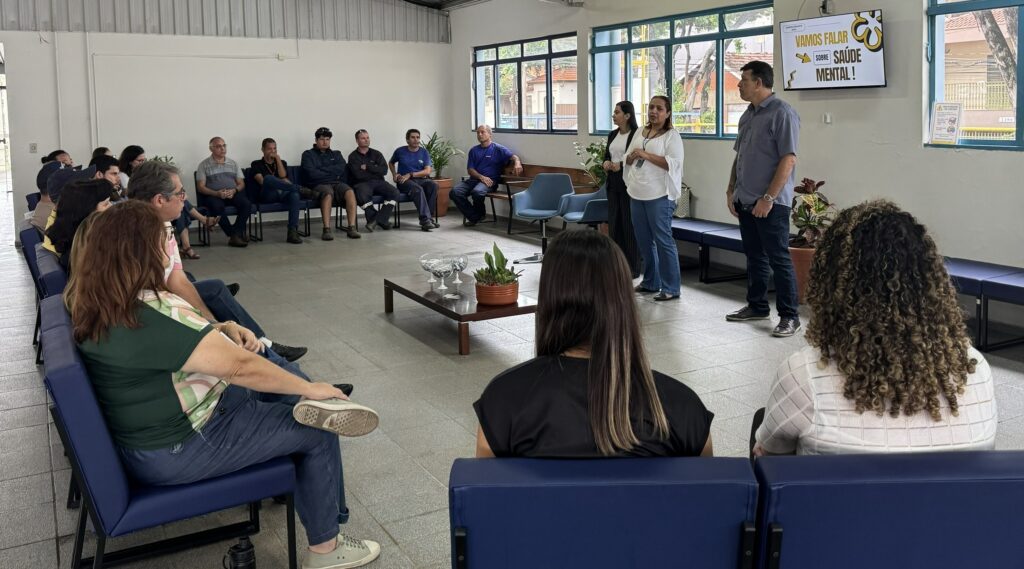 A imagem mostra uma reunião ou palestra sobre saúde mental, realizada em um ambiente interno, possivelmente um escritório ou sala de treinamento. Um grupo de pessoas está sentado em cadeiras e bancos dispostos em semicírculo, prestando atenção a três apresentadores que estão em pé próximos a uma televisão de parede. A televisão exibe a frase "JANEIRO BRANCO 2024 VAMOS FALAR SOBRE SAÚDE MENTAL!", indicando que o tema do encontro é a conscientização sobre saúde mental, associado à campanha Janeiro Branco, que ocorre no início do ano para promover o cuidado com a saúde emocional. O ambiente é claro e organizado, com paredes brancas, piso de cerâmica cinza e janelas amplas que deixam entrar luz natural. No centro da sala há uma mesa baixa de madeira com alguns objetos decorativos, como um vaso com planta. As pessoas assistindo parecem atentas e engajadas, sugerindo um clima de interesse e respeito sobre o assunto tratado. A imagem transmite a importância do diálogo e da informação sobre saúde mental no ambiente de trabalho ou em grupos comunitários.