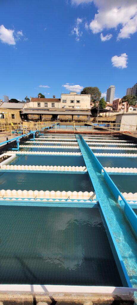 A imagem mostra uma vista aérea de uma estação de tratamento de água. * O céu azul com algumas nuvens brancas está no topo da imagem. * Abaixo do céu, há uma fileira de edifícios de cor clara, alguns com telhados vermelhos. * A maior parte da imagem é ocupada por tanques de água interconectados. Esses tanques são longos e estreitos, com bordas brancas e azuis. * A água nos tanques é de cor verde-escura e parece estar em movimento, com reflexos do céu e das estruturas circundantes. * Uma calha azul atravessa a imagem diagonalmente, transportando água de um tanque para outro. * Em segundo plano, há mais edifícios, incluindo alguns edifícios altos, sugerindo que a estação de tratamento está localizada em uma área urbana. * A iluminação geral sugere que é um dia ensolarado.