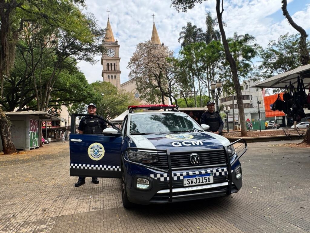 A imagem mostra dois guardas municipais em frente a um carro oficial da GCM (Guarda Civil Municipal). O carro é um Volkswagen T-Cross, de cor azul e branca, com distintivos da GCM e a placa "SWJ1156". Um dos guardas está com a porta do motorista aberta, em pé ao lado do veículo, enquanto o outro está em pé atrás do carro, do lado do passageiro. Ao fundo, observa-se uma praça com árvores frondosas e uma igreja com torre e relógio. Há também algumas barracas de comércio e prédios ao redor, indicando um ambiente urbano. O céu está parcialmente nublado. A cena parece transmitir uma sensação de segurança e presença policial no local.