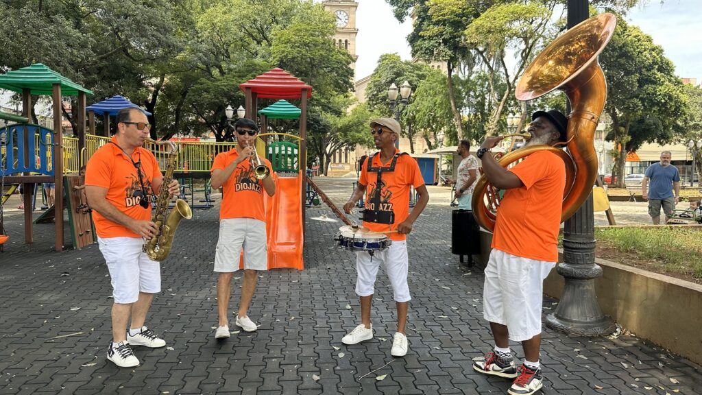 A banda DioJazz se apresenta em um parque. Quatro músicos, vestidos com camisetas laranjas e calças brancas, tocam instrumentos de sopro e percussão. À esquerda, um homem toca um saxofone, enquanto ao seu lado, outro músico toca uma trombeta. Mais ao centro, um terceiro integrante dedilha uma caixa de ritmos presa ao corpo, usando baquetas. Finalizando o quarteto, à direita, um homem com um chapéu preto toca um tuba, um instrumento de sopro grande e brilhante. Ao fundo, uma estrutura de playground colorida e um relógio em uma torre sugerem que a apresentação ocorre em uma praça pública. Outras pessoas podem ser vistas ao fundo, algumas observando a performance.