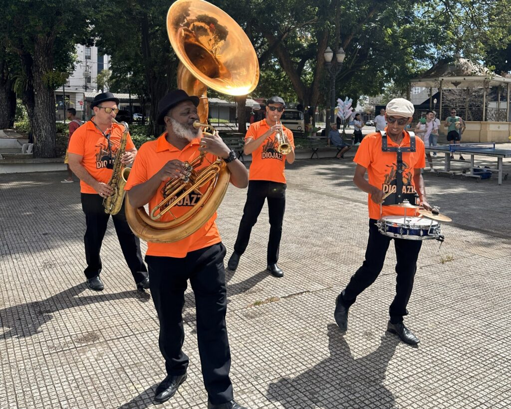 A imagem mostra um grupo de quatro músicos de jazz em um parque. Todos eles usam camisetas laranja com a inscrição "DIOJAZZ" e calças pretas. O músico à esquerda está tocando um saxofone tenor. Ele usa um chapéu preto e óculos escuros. O músico central, mais proeminente, está tocando um sousafone. Ele tem barba grisalha e usa um chapéu preto. O sousafone é um instrumento de bronze grande e curvo. Atrás do músico do sousafone, um músico toca uma trombeta. Ele usa um boné e óculos escuros. O músico mais à direita está tocando uma caixa com baquetas. Ele usa um boné branco e óculos escuros. Ele também está usando um colete com suportes para as baquetas. O fundo da imagem mostra árvores, um coreto, algumas pessoas sentadas e uma mesa de pingue-pongue. O chão é pavimentado com um padrão de ladrilhos. A luz do sol projeta sombras nítidas no chão.