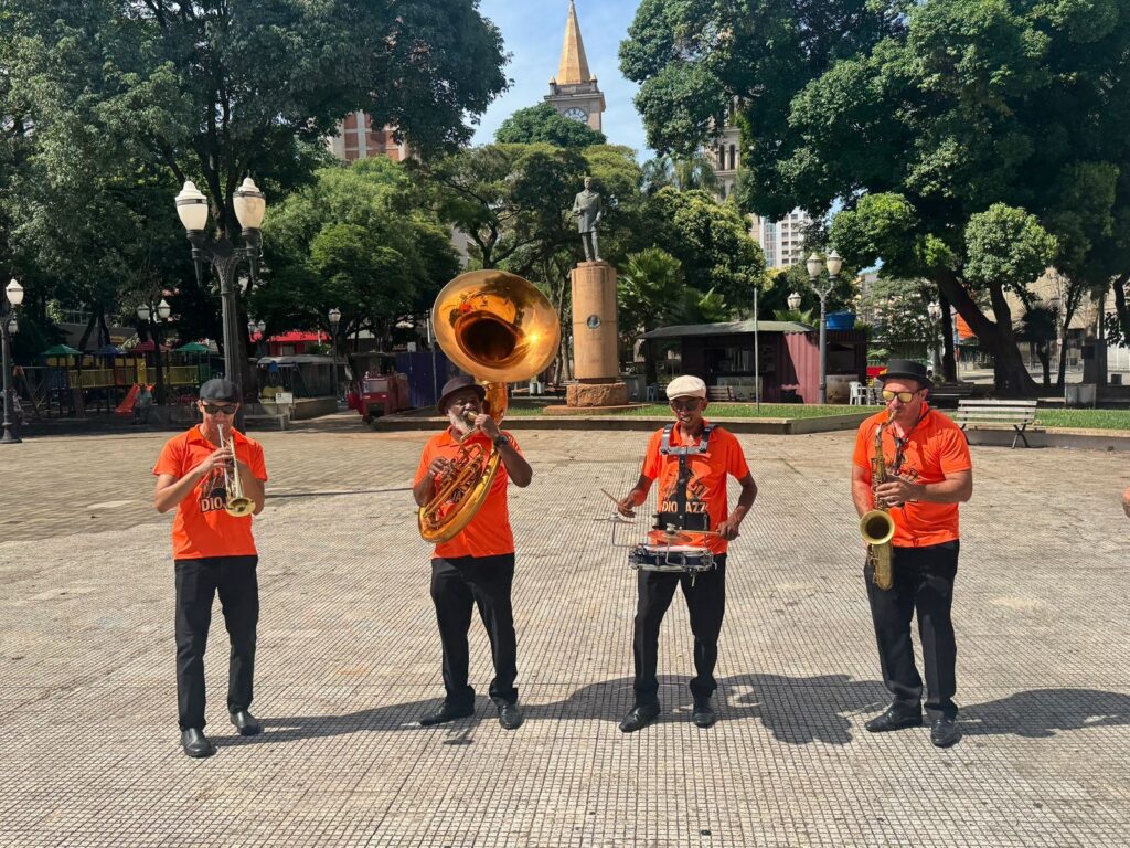 A banda de jazz, vestida com camisas laranja vibrantes, toca em uma praça ensolarada. O músico da esquerda toca trompete, o do centro toca tuba e bateria, e o da direita toca saxofone. Ao fundo, uma estátua em um pedestal alto se destaca contra o céu azul, cercada por árvores frondosas e edifícios históricos. A praça tem um piso quadriculado e um poste de luz ornamentado contribui para o charme da cena.