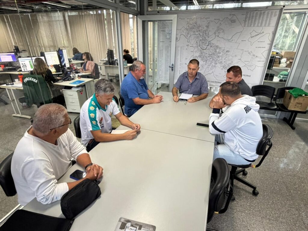 A imagem mostra um grupo de homens reunidos em torno de uma mesa em um escritório. Ao fundo, há várias mesas com computadores onde algumas mulheres trabalham. Há também um grande mapa fixado na parede. Um dos homens sentados à mesa principal está usando um moletom branco com a palavra "RIO" escrita nas costas. Outro homem usa uma camisa polo azul, e um terceiro veste uma camisa polo branca com detalhes verdes. Um quarto homem, com óculos, está de frente para a câmera, enquanto um quinto homem está mais afastado, de costas para a câmera. A atmosfera parece ser de uma reunião de trabalho.