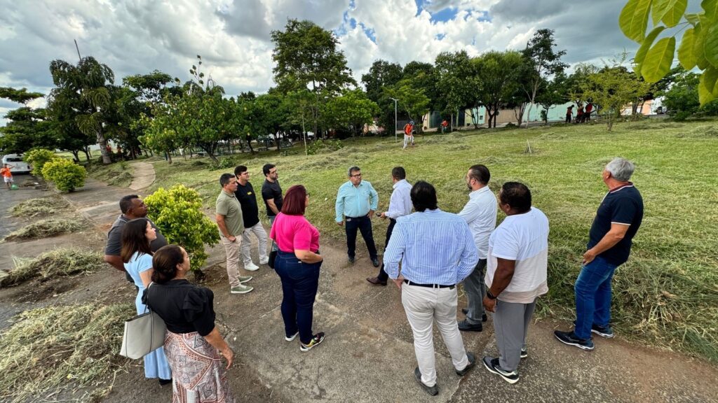 A imagem mostra um grupo de pessoas reunidas em um círculo em um parque ou área verde. A maioria das pessoas está vestida de forma casual, com alguns homens usando camisas sociais e calças, e algumas mulheres usando vestidos e calças. O cenário é um gramado com árvores ao redor e um céu nublado. Há também algumas pilhas de grama cortada ao longo do caminho de concreto onde o grupo está reunido. No fundo, é possível ver algumas pessoas trabalhando na grama e um carro estacionado. A atmosfera parece ser de uma reunião informal ou uma inspeção do local.