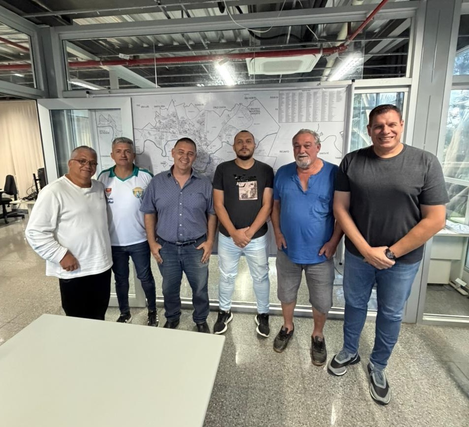 A imagem mostra um grupo de seis homens em pé em uma sala, possivelmente um escritório, com um grande mapa ou diagrama na parede atrás deles. Eles estão todos vestidos de forma casual. Do lado esquerdo, um homem mais velho com óculos usa uma camisa branca de manga comprida e calças pretas. Ao lado dele, um homem mais jovem veste uma camisa polo branca com detalhes verdes e calças jeans. O terceiro homem da esquerda para a direita usa uma camisa xadrez azul e jeans. No centro da imagem, um homem com barba usa uma camiseta preta com uma estampa gráfica e jeans rasgados. Ao seu lado, um homem mais velho com barba grisalha veste uma camiseta azul com decote em V e shorts cinza. O homem mais à direita da imagem usa uma camiseta cinza escura e jeans. A sala parece ter um teto exposto com tubulações e iluminação. Há também uma grande mesa branca na frente do grupo, sugerindo que a foto foi tirada em um ambiente de reunião ou escritório.