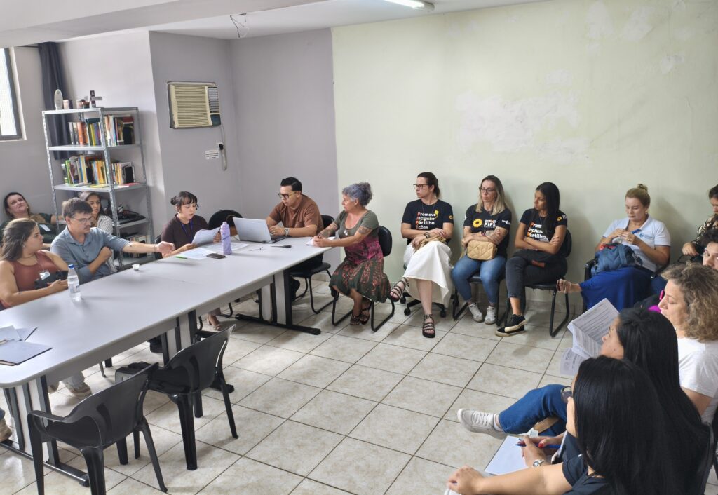 A imagem mostra um grupo de pessoas reunidas em uma sala, possivelmente para uma reunião ou evento. No primeiro plano, à esquerda, uma mulher com uma blusa marrom está sentada à mesa, olhando para a frente. Ao lado dela, um homem de camisa azul está sentado, gesticulando com a mão enquanto conversa com alguém. Uma garrafa de água está sobre a mesa. A mesa longa domina a parte inferior da imagem, com várias pessoas sentadas ao redor dela. Algumas estão olhando para papéis, outras para um laptop aberto. Ao fundo, à esquerda, há uma estante de livros cheia e um ar condicionado na parede. No lado direito da sala, um grupo de pessoas está sentado em cadeiras, algumas em um semicírculo. Muitas delas estão usando camisetas pretas com texto, e algumas carregam bolsas. Uma mulher de camisa branca está sentada mais afastada, parecendo tricotar.