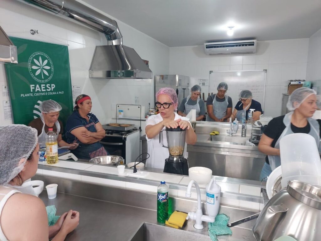 A imagem mostra um grupo de pessoas em uma cozinha industrial ou laboratório culinário, provavelmente participando de um curso ou workshop prático. No centro da imagem, uma mulher com touca rosa, óculos e avental branco está utilizando um liquidificador que contém amendoins ou algum tipo de grão. Ao redor dela, outras pessoas, também com toucas e aventais, observam atentamente a atividade. O ambiente é limpo e organizado, com paredes revestidas de azulejos brancos e uma coifa industrial acima da bancada. Há uma bandeira verde ao fundo com o logotipo da FAESP (Federação da Agricultura e Pecuária do Estado de São Paulo) e o slogan "Plante, cultive e colha a paz!". Sobre a bancada, há diversos itens, como uma garrafa de óleo, um frasco de detergente da marca Ypê e utensílios de cozinha. O cenário sugere uma aula prática voltada para técnicas agrícolas ou culinárias, promovida por uma entidade ligada à agricultura e pecuária do estado de São Paulo. As expressões das pessoas indicam concentração e interesse na atividade realizada.