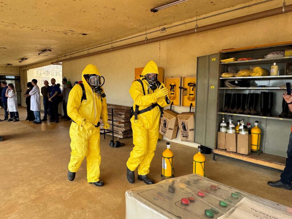 A imagem mostra duas pessoas vestindo trajes de proteção química amarelos completos, incluindo capuzes, máscaras de gás com filtros e luvas. Elas estão em um ambiente interno com piso de concreto e paredes pintadas de amarelo. À esquerda, há um grupo de pessoas em roupas casuais, observando. Há também prateleiras com equipamentos e caixas. No centro e à direita, vemos equipamentos de proteção, como botas de borracha em uma prateleira metálica, cilindros de gás amarelos e caixas de papelão com a marca "Dräger PA 540". Há também painéis de controle com botões coloridos em primeiro plano. O ambiente parece ser um local de treinamento ou armazenamento para situações de risco.