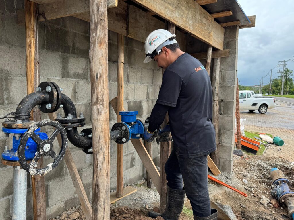 A imagem mostra um trabalhador de capacete branco com o logo "SANE SOLUTI" trabalhando em uma instalação hidráulica. Ele veste uma camiseta preta com o mesmo logo nas costas, calças pretas e botas de borracha. O trabalhador está concentrado em apertar uma conexão em um tubo azul com uma chave inglesa. Ao redor, há uma estrutura de madeira improvisada sustentando vigas de madeira e blocos de concreto. A instalação hidráulica inclui tubos pretos curvos com flanges e um medidor azul com uma pequena caixa preta em cima. Há também uma válvula grande com uma roda preta. No fundo, ao lado de uma estrada, um caminhão branco está estacionado. O céu está nublado, indicando um dia chuvoso ou com previsão de chuva. O ambiente parece ser um canteiro de obras.
