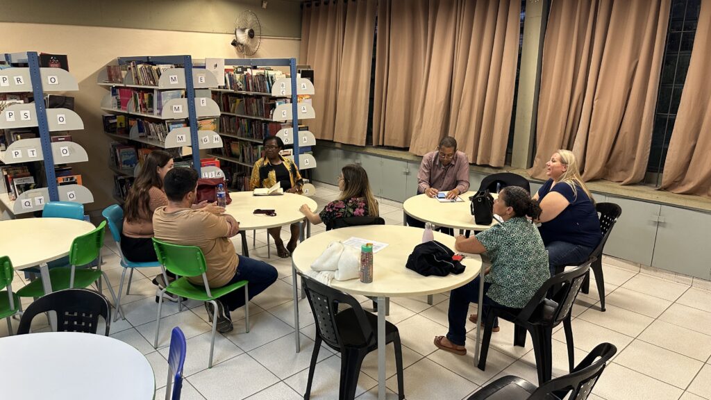 A imagem mostra um grupo de pessoas sentadas em torno de mesas redondas em uma biblioteca. Há estantes de livros ao fundo, algumas com etiquetas de letras visíveis. As pessoas parecem estar em uma reunião ou discussão. Uma mulher está sentada à esquerda, com um livro aberto à sua frente. Um homem está sentado em frente a ela. Outras pessoas estão sentadas em torno de outras mesas, conversando. As cortinas nas janelas ao fundo são de cor bege. O chão é revestido com azulejos brancos. Há um ventilador de teto na parede acima das estantes de livros.