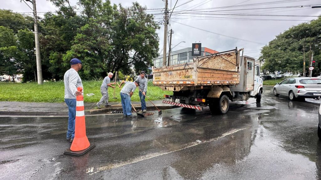 A imagem mostra uma cena urbana durante um dia chuvoso ou logo após a chuva, com o chão molhado refletindo a luz difusa do céu nublado. Quatro trabalhadores estão realizando a limpeza de uma boca de lobo (ralo de drenagem) na calçada, usando pás e vassouras para remover detritos. Eles vestem roupas de trabalho simples, como camisetas e calças jeans. Ao centro-direita da imagem, há um caminhão branco com a caçamba enferrujada, provavelmente usado para recolher o lixo retirado do bueiro. À esquerda, um cone laranja sinaliza a área de trabalho, e um dos trabalhadores está de pé próximo a ele, possivelmente coordenando ou observando. Ao fundo, vê-se uma área verde com árvores, algumas construções e uma placa que indica "PAN BIANCO ACADEMIA", sugerindo o local na cidade de Piracicaba-SP. Um carro prateado está estacionado ou passando à direita da imagem. A cena transmite uma rotina de serviço público de manutenção urbana, com foco na limpeza e prevenção de alagamentos, em um ambiente típico de cidade brasileira.