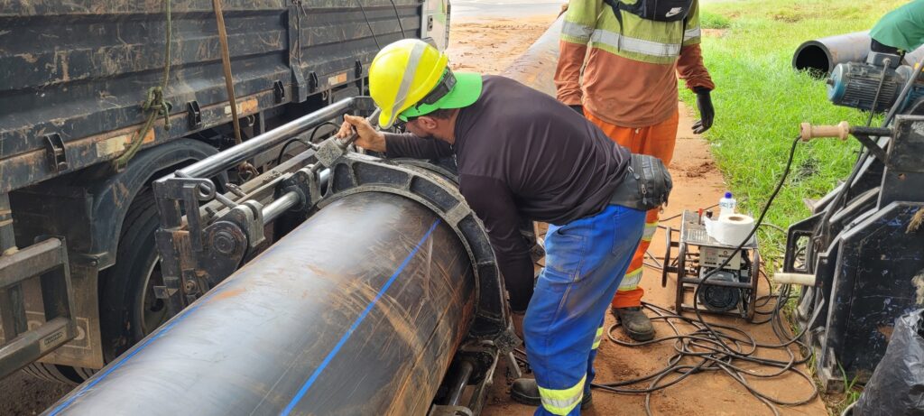 A imagem mostra dois trabalhadores em um canteiro de obras, possivelmente realizando a instalação de um grande tubo. Um trabalhador, vestindo um capacete amarelo e um boné verde, está debruçado sobre o tubo, que parece ser de plástico preto com duas linhas azuis. Ele usa um macacão azul e um cinto de ferramentas. O outro trabalhador, ao fundo, veste um colete de alta visibilidade laranja e verde, e luvas pretas. O tubo está apoiado em uma estrutura metálica robusta, possivelmente parte de um equipamento de soldagem ou junção de tubos, que está conectado a um gerador elétrico. Há cabos espalhados pelo chão, e um rolo de papel higiênico e uma garrafa de água estão sobre o gerador. Ao lado, um caminhão com a lateral suja e enferrujada sugere que o material foi transportado para o local. A área ao redor é de terra batida e grama, indicando um ambiente de construção externa.