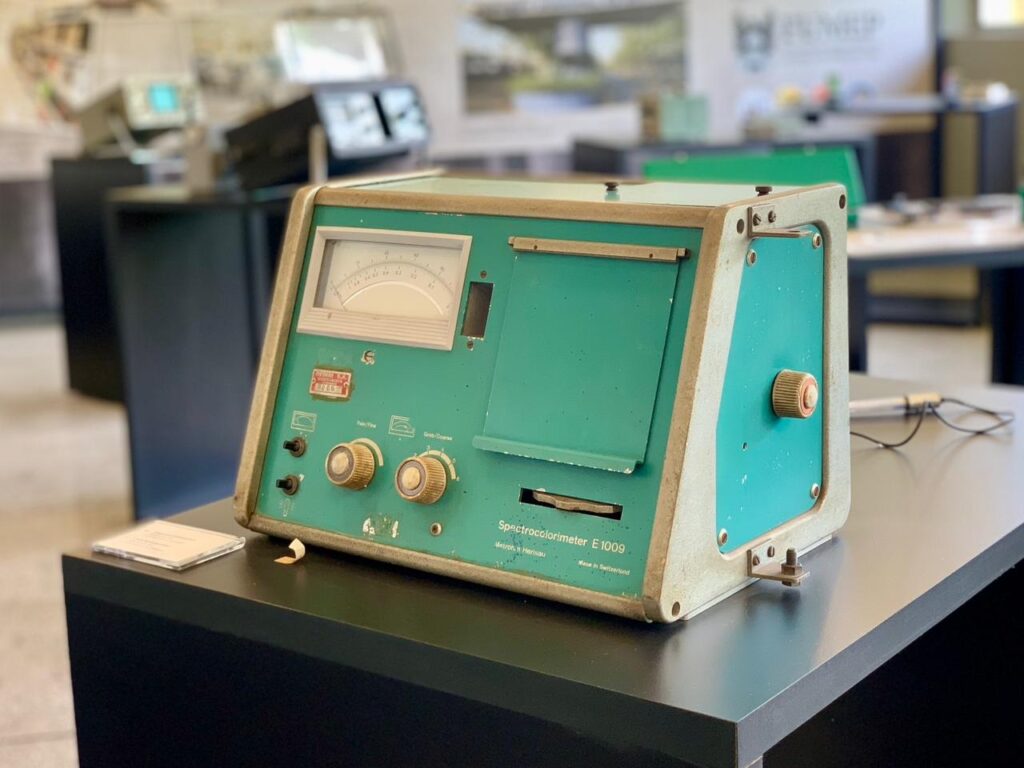 Na imagem, um espectrocolorímetro E 1009, um instrumento científico de cor verde e cinza, está posicionado sobre uma mesa preta. O aparelho possui um mostrador analógico circular na parte superior esquerda, com uma agulha indicando valores em uma escala. Abaixo do mostrador, há vários botões e seletores, incluindo um seletor rotativo com as marcações "Fine/Fine" e "Grad/Coarse". Há também uma abertura retangular na parte frontal do instrumento, possivelmente para a inserção de amostras. Uma etiqueta com o texto "Spectrocolorimeter E 1009", "G.B.H. Herisau" e "Made in Switzerland" está visível na parte inferior frontal do aparelho. Um pequeno cartão branco está posicionado ao lado esquerdo do instrumento. O fundo da imagem mostra um ambiente interno, com outras mesas, equipamentos eletrônicos e um logotipo desfocado em uma parede, sugerindo um laboratório ou museu de ciência.