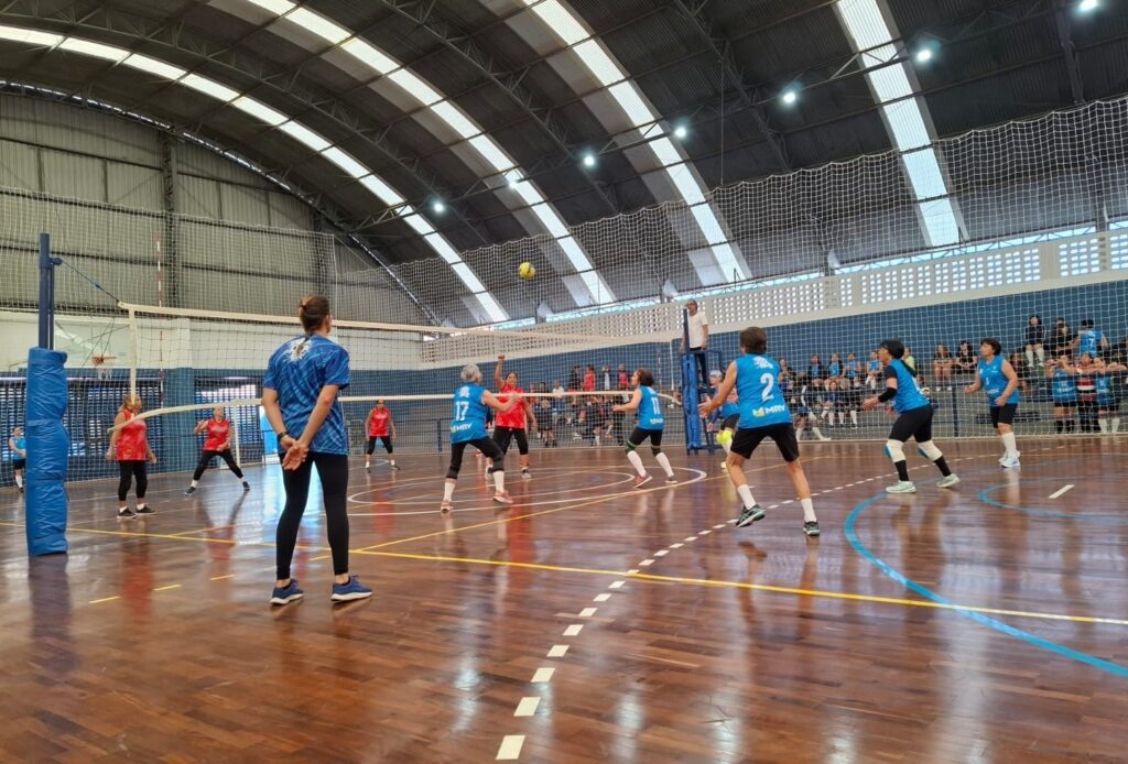 A imagem mostra um jogo de vôlei em uma quadra interna com piso de madeira. Há uma rede dividindo a quadra, com jogadoras em ambos os lados. As jogadoras vestem uniformes de cores diferentes, algumas de azul e outras de vermelho. Uma bola de vôlei está no ar, acima da rede. Há também um árbitro em uma plataforma alta ao lado da rede. Ao fundo, espectadores estão sentados nas arquibancadas. A iluminação vem de cima, com luminárias no teto da quadra.
