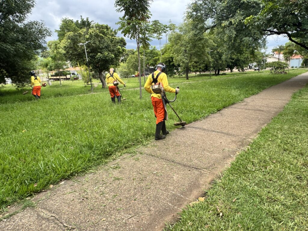 A imagem mostra três homens uniformizados com coletes amarelos e calças laranjas, aparando a grama com roçadeiras em um parque ou área verde. Eles estão em uma linha, cada um trabalhando em sua seção da grama, com o caminho de concreto passando ao lado. O dia parece nublado, mas a vegetação está exuberante e verde. Em segundo plano, há árvores, postes de luz e alguns veículos.