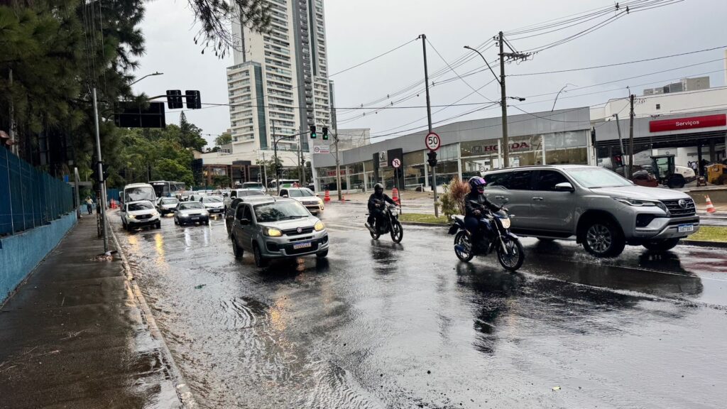 A imagem mostra uma rua movimentada de uma cidade brasileira em um dia chuvoso, com o asfalto molhado e reflexos evidentes na pista. No primeiro plano, há vários veículos, incluindo um Fiat Mobi prata com a placa "CUN5C47" e um Toyota SW4 prata, além de duas motocicletas com os pilotos usando capacetes e roupas escuras. Ao fundo, vê-se prédios comerciais, entre eles uma concessionária Honda, e um edifício residencial alto e branco. A rua está sinalizada com placas de limite de velocidade de 50 km/h, semáforos e postes com muitos fios elétricos. À esquerda, há uma calçada com muro azul e árvores do tipo pinheiro. O clima é típico de uma cidade em um dia nublado e chuvoso, transmitindo uma sensação de movimento e rotina urbana sob a chuva.