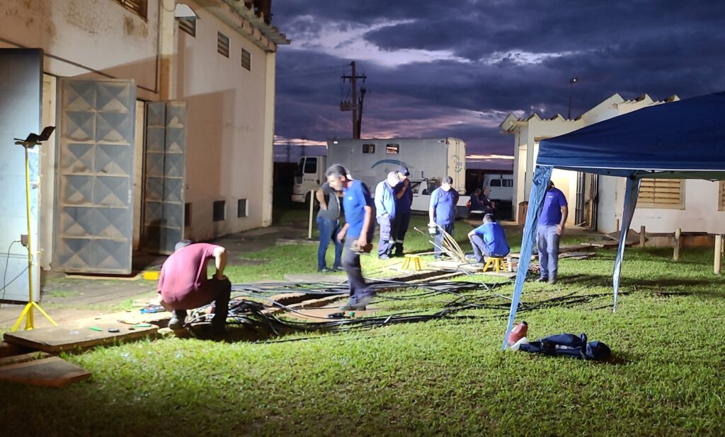 A imagem mostra um grupo de pessoas em um local externo, possivelmente uma área de serviço ou industrial, durante o crepúsculo. Há uma construção ao fundo com grandes portas metálicas e janelas. Várias pessoas vestindo uniformes azuis e um indivíduo com uma camisa vinho estão reunidos em torno de uma área gramada onde há muitos cabos e equipamentos elétricos. Algumas pessoas parecem estar trabalhando ou inspecionando os cabos, enquanto outras observam. Há também uma tenda azul montada, e ao fundo, veículos e um contêiner. A iluminação artificial, proveniente de um foco de luz amarelo à esquerda, ilumina a área de trabalho, contrastando com o céu escuro e nublado.