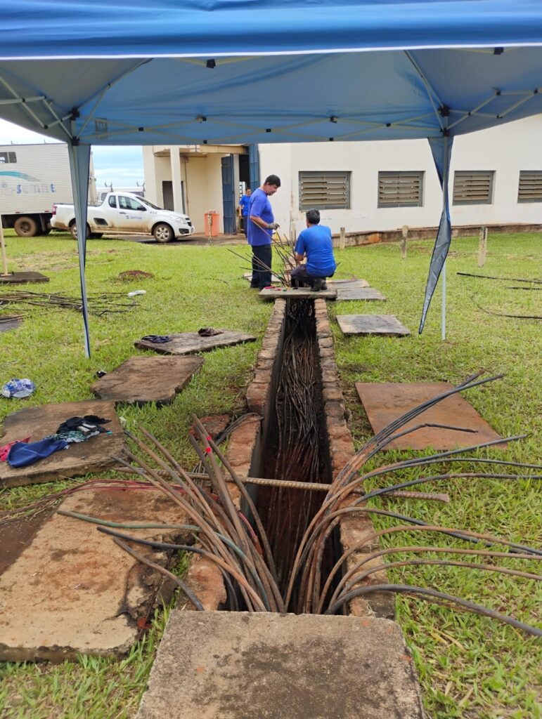 * Dois homens com camisas azuis estão trabalhando ao ar livre sob uma tenda azul. * Eles estão posicionados perto de uma vala aberta cheia de vários cabos grossos. * Um homem está em pé segurando alguns cabos, enquanto o outro está de joelhos e parece estar organizando ou conectando-os. * A vala é revestida com tijolos, e várias lajes de concreto estão espalhadas pela área gramada. * Ao fundo, há um prédio com janelas e uma caminhonete branca com o logo "semae". * A cena sugere uma tarefa de manutenção ou instalação de infraestrutura envolvendo cabos elétricos ou de comunicação.