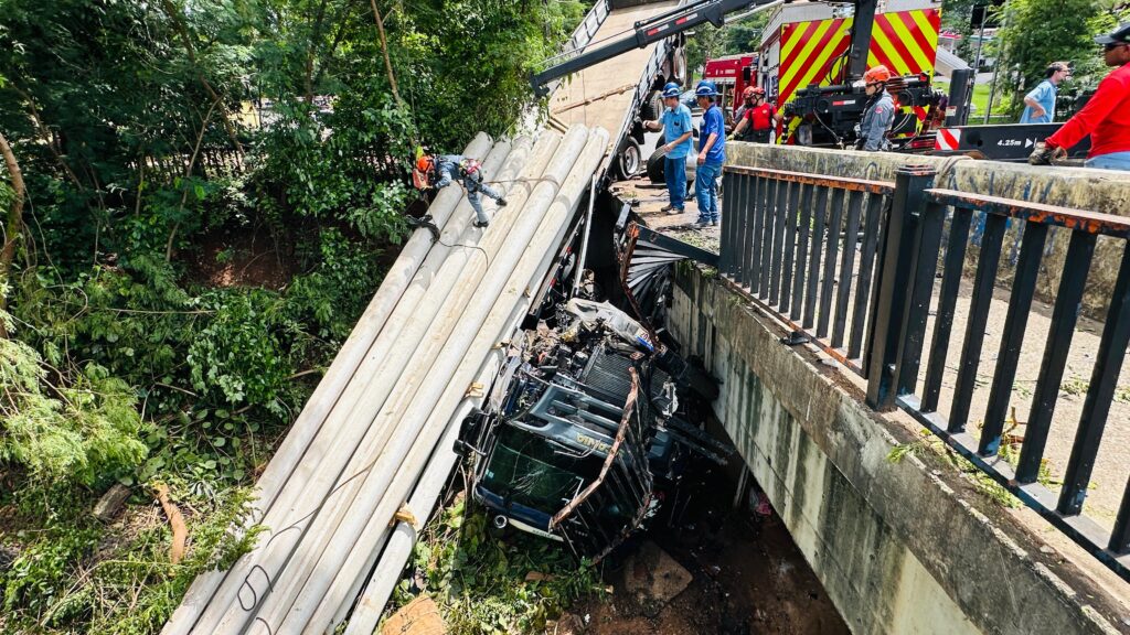 A imagem mostra as consequências de um acidente em uma ponte, onde um caminhão carregado de postes de concreto tombou. A carga se espalhou pela ponte e parte dela caiu em um barranco coberto de vegetação. Uma equipe de resgate está no local, com pessoas usando capacetes e coletes de segurança. Um bombeiro está descendo com equipamentos de rapel sobre os postes caídos, avaliando a situação. Ao fundo, um caminhão guindaste e um caminhão de bombeiros estão posicionados na ponte, indicando a operação de resgate e remoção dos destroços. Pessoas em roupas civis também observam a cena. A ponte possui uma mureta com grades de proteção, que parece ter resistido ao impacto. A vegetação ao redor sugere que o local é uma área arborizada.