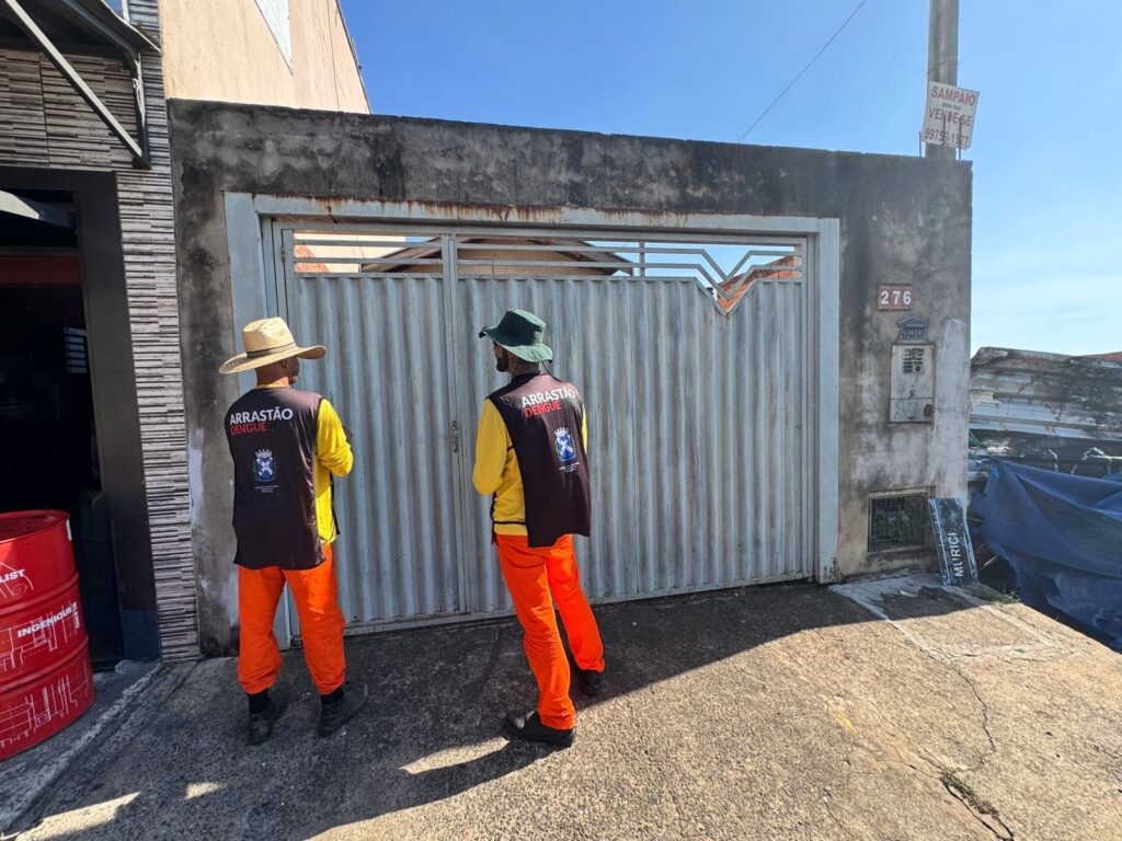 A imagem mostra dois homens vestindo coletes com a inscrição "ARRASTÃO DENGUE 2023" e chapéus, um de palha e outro de tecido verde. Eles estão em frente a um portão metálico cinza, que parece estar danificado em algumas partes. As calças dos homens são de cor laranja vibrante, e eles usam sapatos pretos. No lado direito da imagem, há um poste com uma placa de "VENDE-SE" e um número de telefone. Abaixo do portão, há uma caixa de correio e uma pequena grade com vegetação. À direita, há uma pilha de materiais cobertos por uma lona azul. No lado esquerdo, há tambores vermelhos empilhados. O céu está azul e claro, indicando um dia ensolarado.