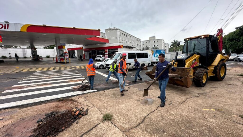 A imagem mostra um grupo de cinco trabalhadores da construção civil realizando reparos em uma via pública. Eles estão usando pás para mover asfalto e detritos na rua. Um dos trabalhadores, em primeiro plano, está caminhando em direção a uma retroescavadeira amarela da marca Caterpillar (CAT), modelo 416E, que está carregada com asfalto em sua concha. A rua possui uma faixa de pedestres pintada em branco. Ao fundo, há um posto de combustível da Shell, com o logotipo visível e os preços dos combustíveis (4,29 e 5,89). Também é possível ver alguns carros estacionados próximos ao posto e edifícios residenciais ou comerciais ao redor. O céu está nublado, sugerindo um dia cinzento. A foto foi tirada de um ângulo levemente elevado, proporcionando uma visão ampla da cena de trabalho.