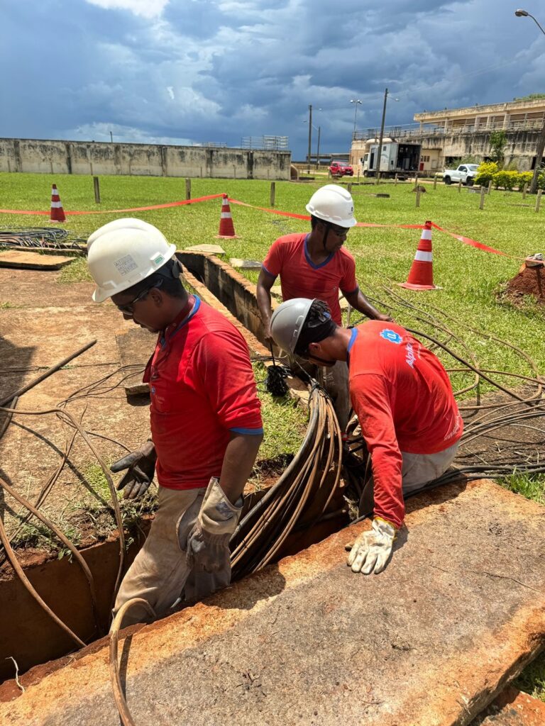 A imagem mostra três trabalhadores em um canteiro de obras, todos usando capacetes e roupas de trabalho vermelhas. Eles estão em torno de uma vala aberta, onde há um grande feixe de cabos grossos. Dois dos trabalhadores estão curvados sobre a vala, parecendo inspecionar ou manipular os cabos. O terceiro trabalhador está um pouco mais atrás, também observando a área. O ambiente parece ser externo, com grama verde e um céu nublado e dramático ao fundo. Há cones de trânsito laranja e branco espalhados pela área, indicando uma zona de trabalho ou perigo. Ao fundo, podem ser vistas algumas construções, incluindo um prédio de vários andares e veículos como caminhões e carros. A cena sugere um trabalho de infraestrutura ou manutenção, possivelmente relacionado a fiação elétrica ou de comunicação.
