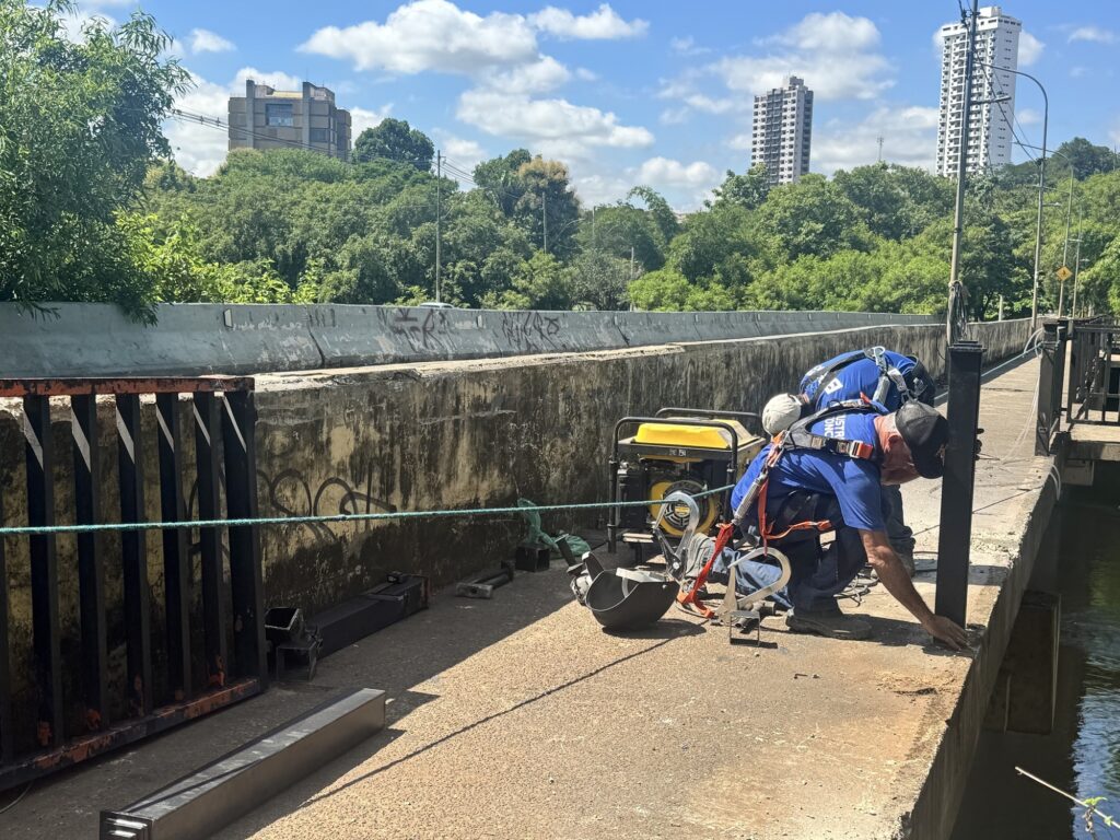 A imagem mostra dois trabalhadores em um dia ensolarado, realizando algum tipo de trabalho em uma estrutura de concreto, possivelmente uma ponte ou um canal. Um gerador amarelo está posicionado perto deles, e há ferramentas e materiais de construção espalhados pela área. Os trabalhadores estão usando equipamentos de segurança, como cintos de segurança. Ao fundo, há uma paisagem urbana com edifícios altos e vegetação exuberante. O céu está azul com algumas nuvens brancas.