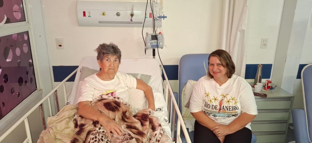 A imagem mostra duas mulheres em um quarto de hospital. Uma mulher mais velha está sentada na cama do hospital, coberta com um cobertor. Ela tem cabelos curtos e grisalhos e está usando uma camiseta branca. Ao lado dela, sentada em uma cadeira, está uma mulher mais jovem com cabelos castanhos. Ela também está usando uma camiseta branca, esta com uma estampa de "Rio de Janeiro" com palmeiras e um carro vintage. O quarto de hospital tem equipamentos médicos visíveis, como um suporte de soro, e uma janela com um padrão circular