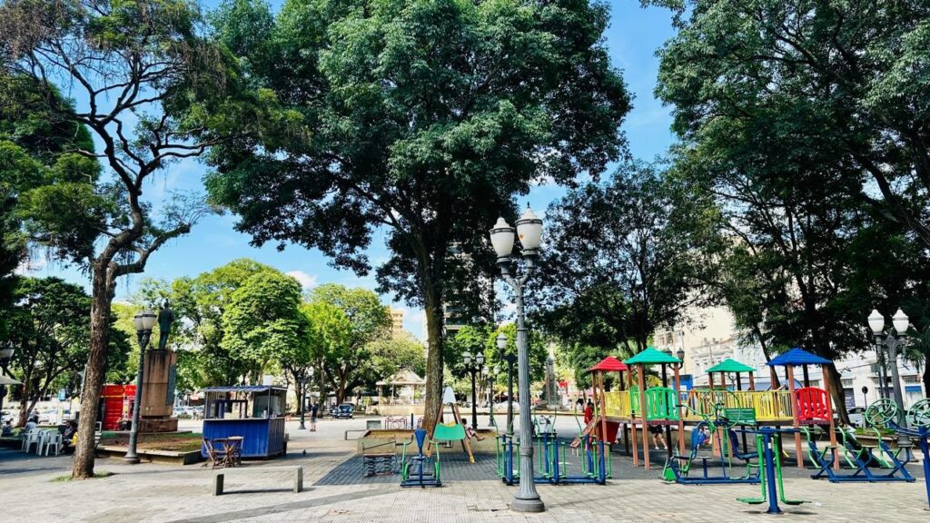 A imagem retrata uma praça pública vibrante, repleta de atividades ao ar livre. No centro, um parque infantil colorido com escorregadores, balanços e estruturas de escalada convida à diversão. Ao redor, equipamentos de ginástica ao ar livre oferecem opções para exercícios físicos. Árvores frondosas e imponentes criam uma atmosfera sombreada e agradável, enquanto bancos e mesas convidam ao descanso e à socialização. Uma pequena barraca azul, possivelmente um quiosque, adiciona um toque pitoresco ao cenário. Ao fundo, edifícios históricos e o céu azul completam a paisagem urbana. A praça parece ser um local movimentado, com pessoas desfrutando do espaço e das atividades disponíveis.